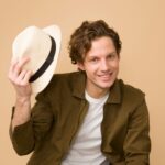 A cheerful man with curly hair tips his hat in a bright studio portrait.